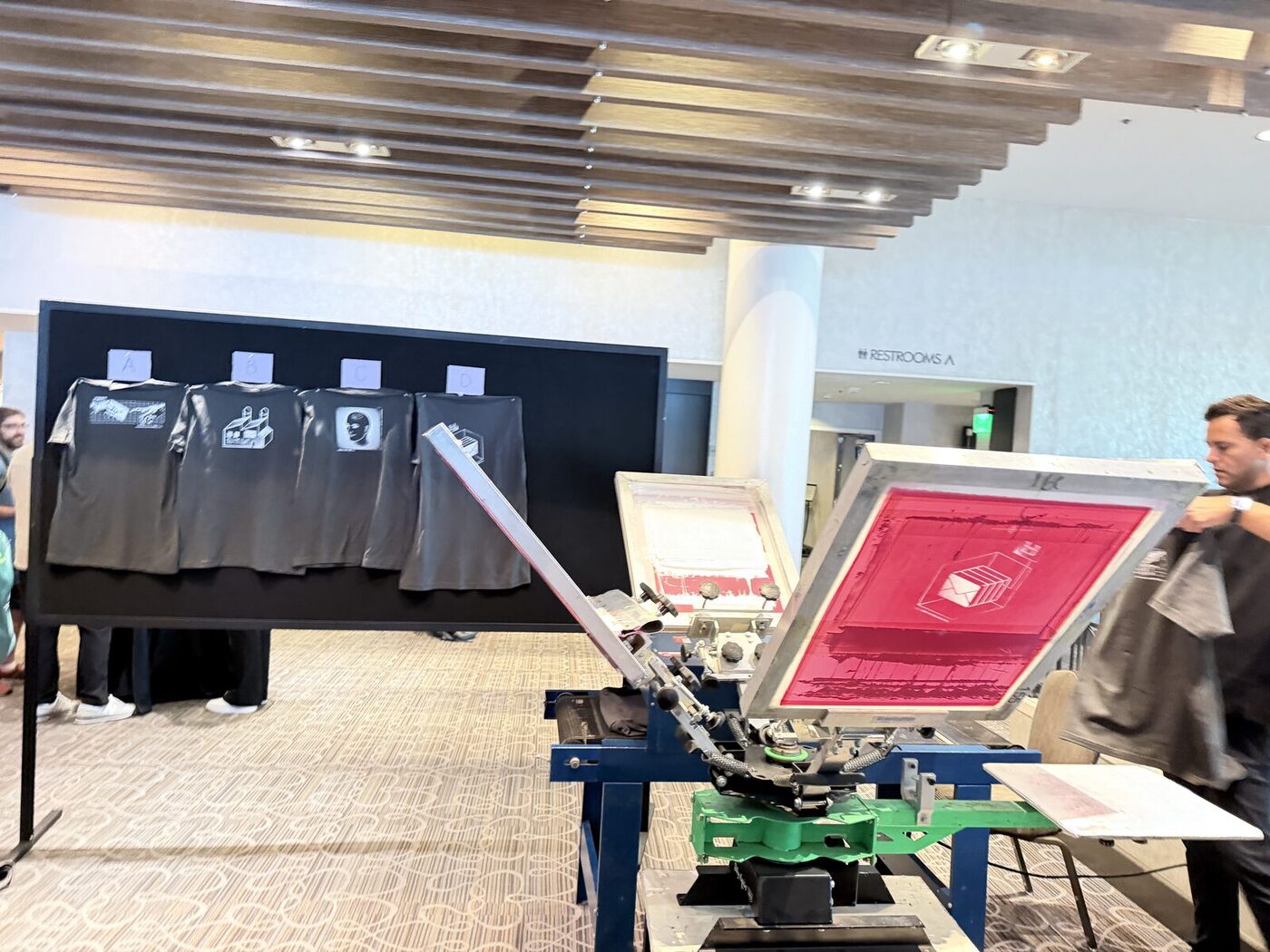 Manual screen-printing press set up at a venue with printed sample shirts displayed behind it